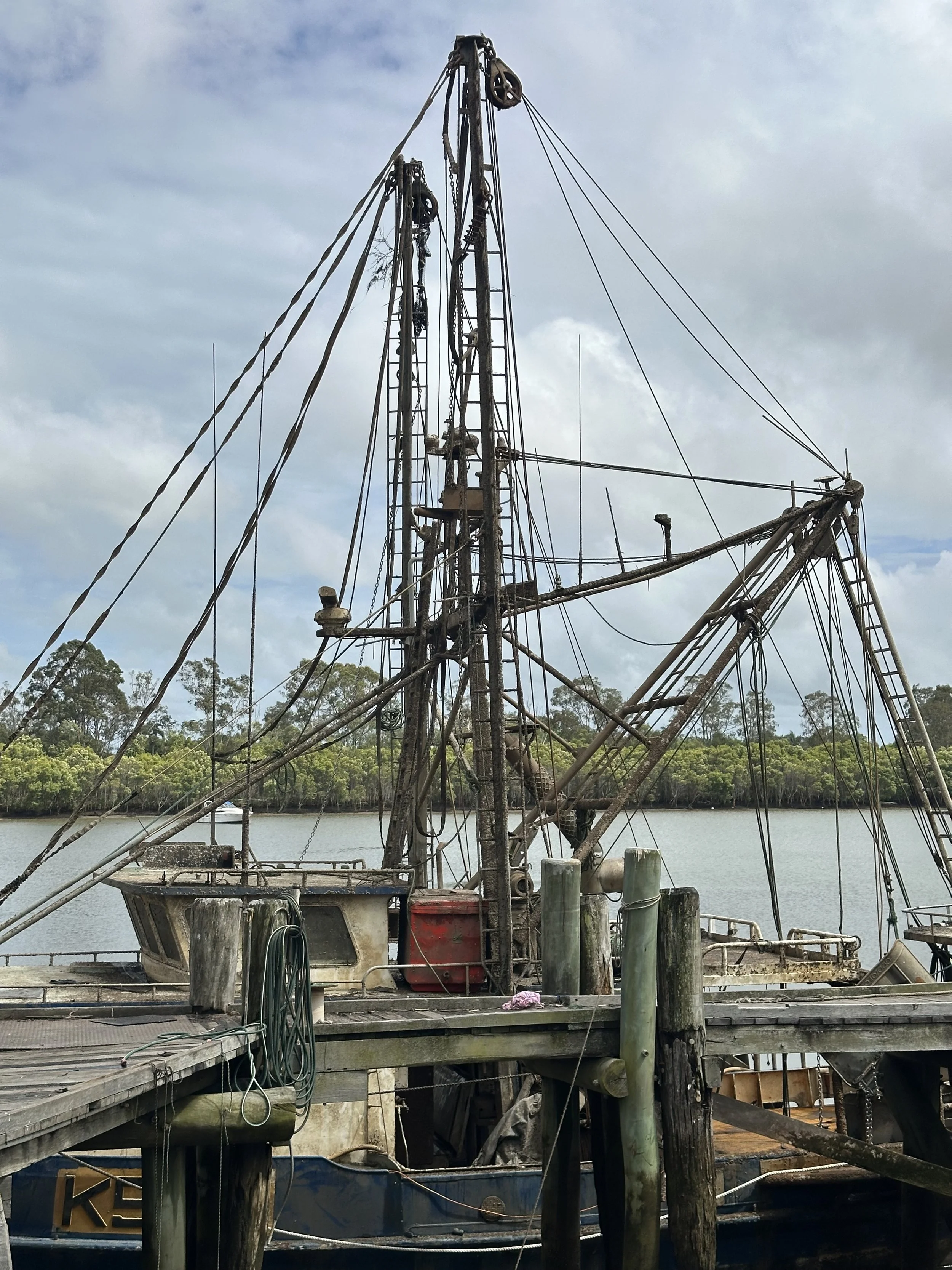 Rusty old boat with tall masts docked at a pier on calm water, with trees and cloudy sky in the background.