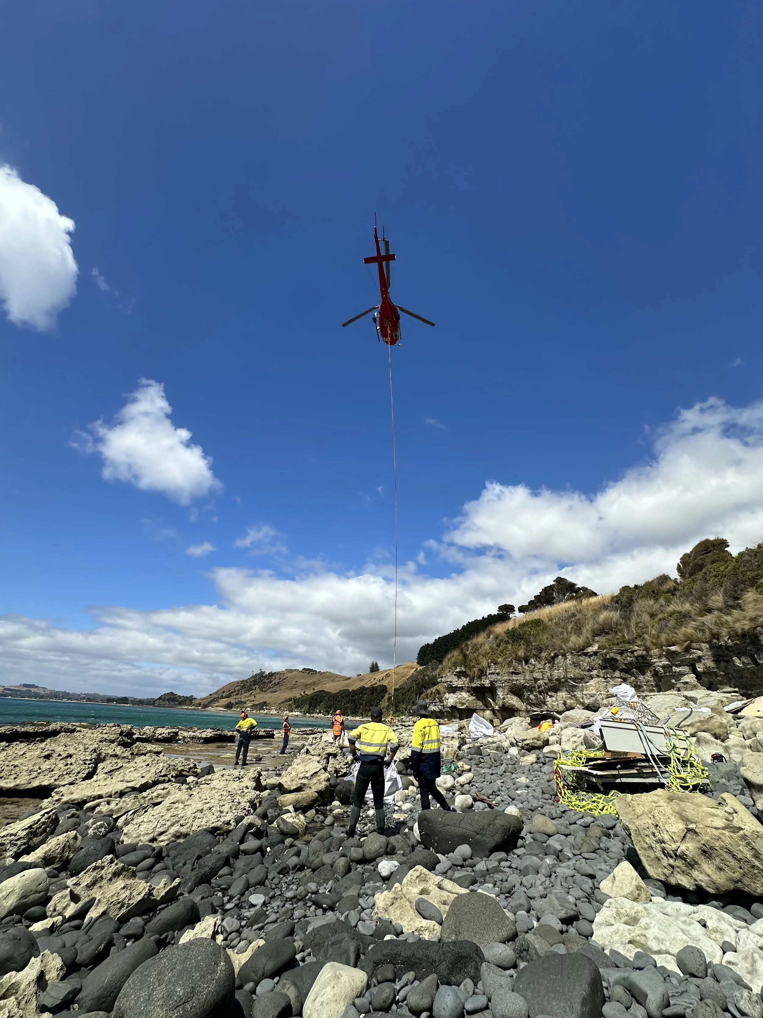 Rescue team on rocky shoreline with helicopter overhead during rescue operation.