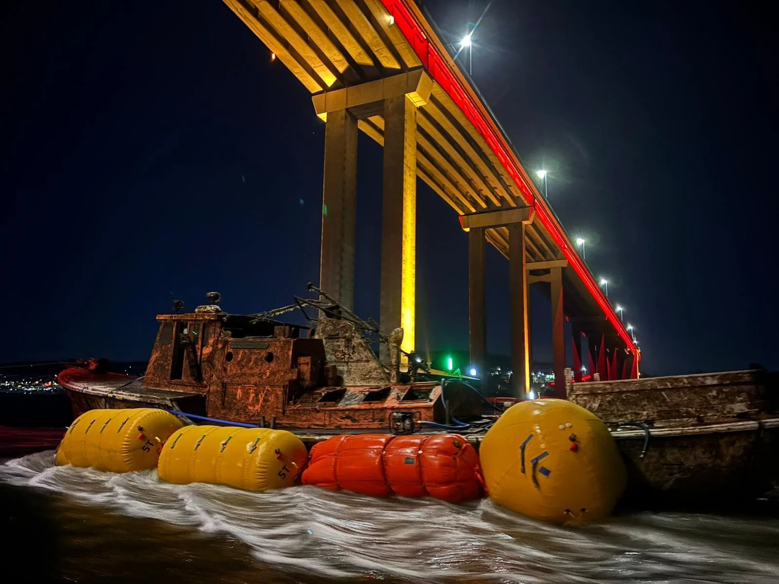 A damaged boat with yellow and orange life buoys floats under a brightly lit bridge at night.