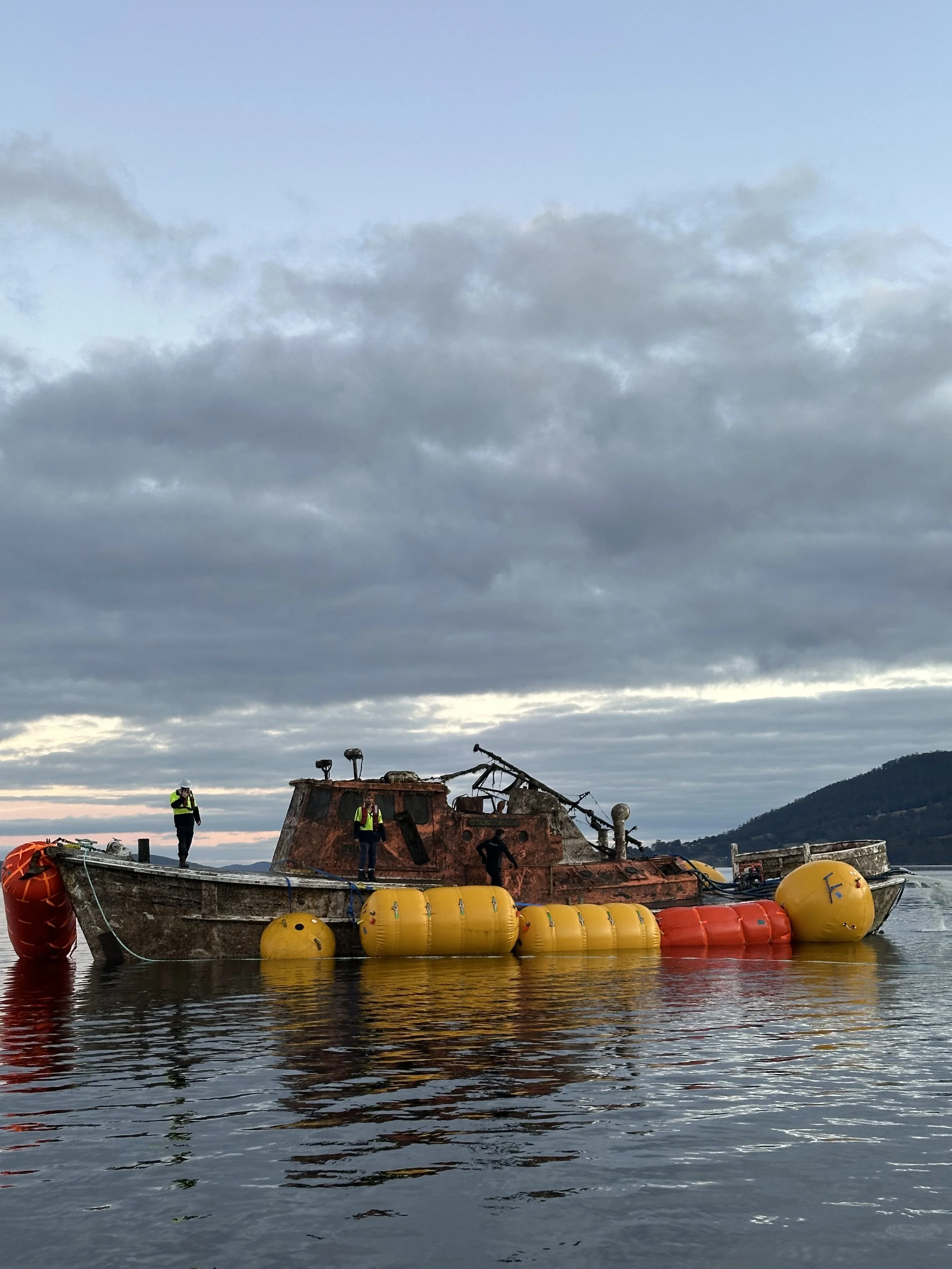 A rusted, abandoned boat with yellow and red flotation devices attached is floating on water. Three people in safety vests are on the boat, with mountains in the background under a cloudy sky.