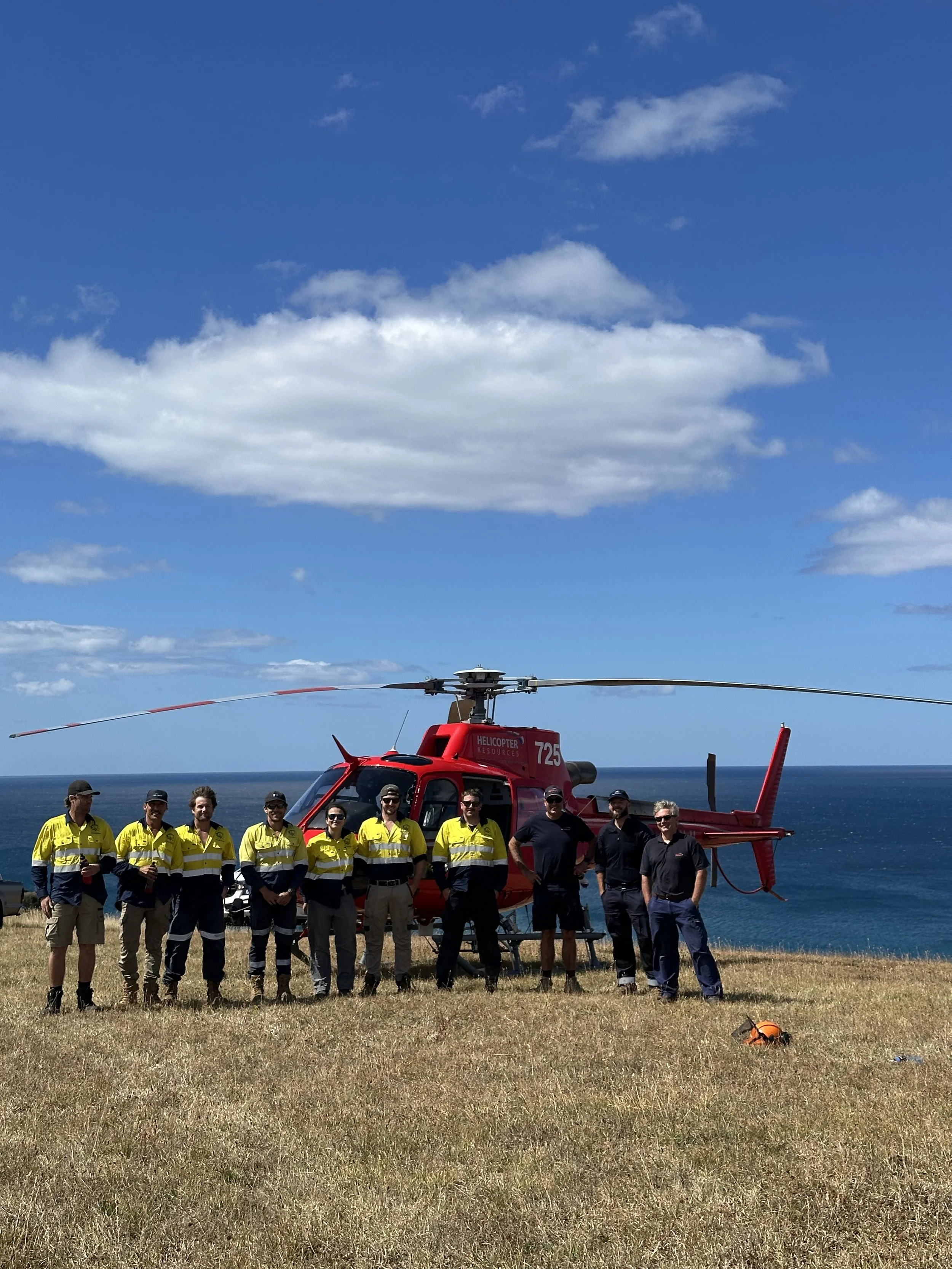 Group of people standing in front of a red rescue helicopter by the coast, with blue sky and ocean in the background.