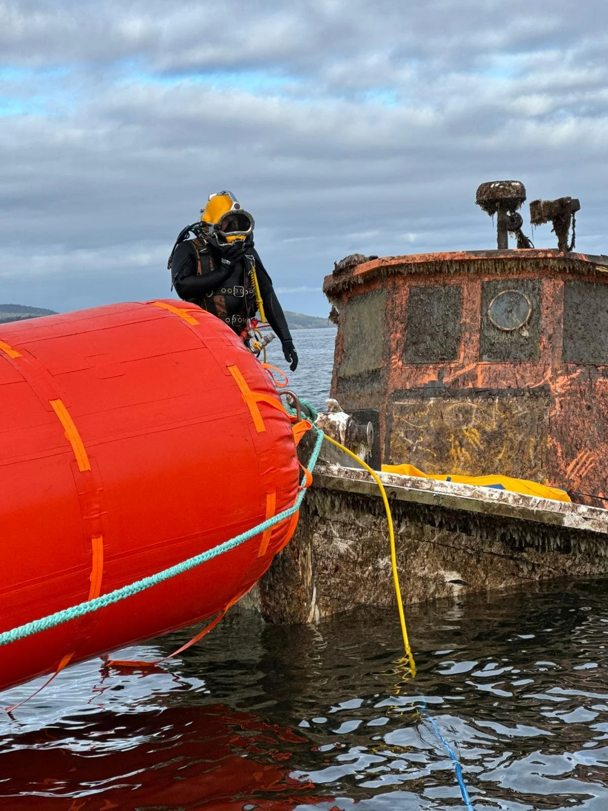A diver in a black wetsuit and yellow helmet standing on a small boat next to a rusty, abandoned shipwreck.