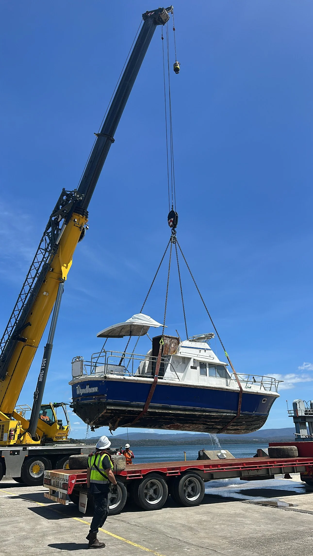 A large yellow crane lifting a boat out of the water onto a truck at a dock. The boat is a white and blue yacht named 'Wyndame' and is suspended by straps from the crane. Workers in safety vests and hard hats are observing the operation near the dock