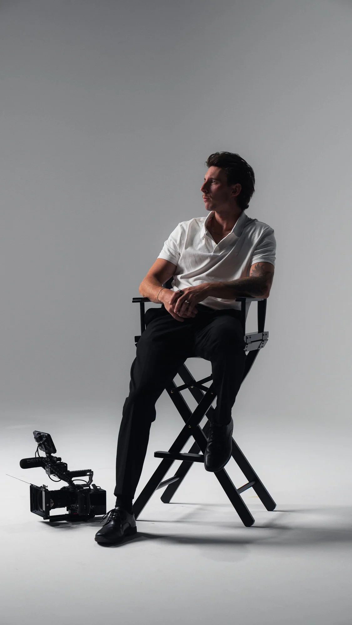 A man sitting on a director's chair in a studio setting, wearing a white shirt and black pants, with a camera on a slider positioned on the floor nearby.