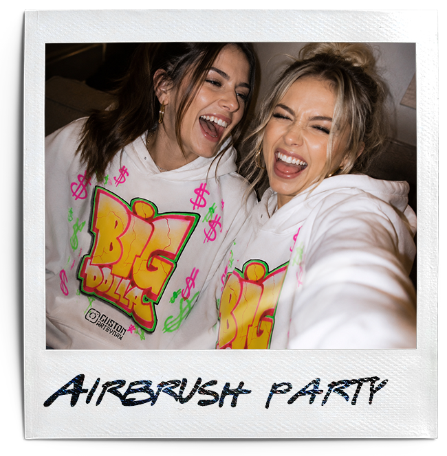 Two women smiling and laughing at an Airbrush Party, wearing matching white hoodies with colorful, graffiti-style designs.