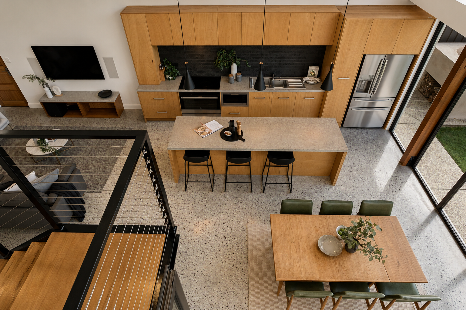 Top-down view of a modern open-concept kitchen and dining area with wooden cabinetry, a kitchen island with black bar stools, a dining table with green chairs, and sliding glass doors leading outside.
