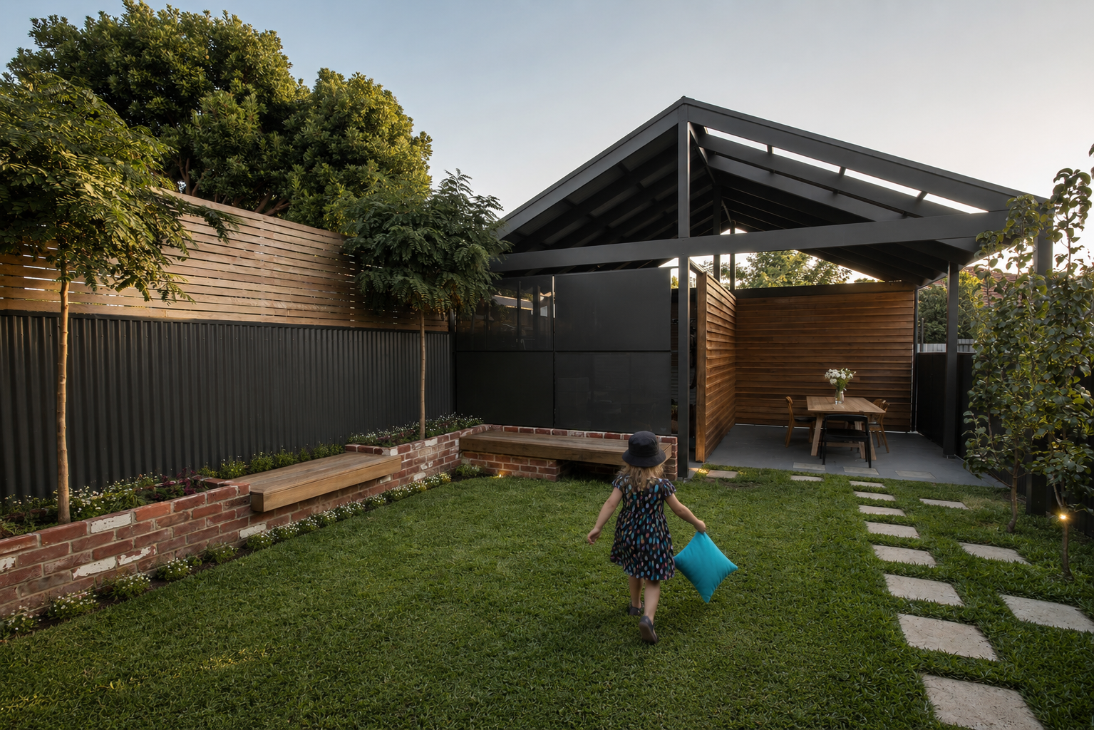 A child wearing a polka dot dress and sun hat running on a green lawn, holding a blue cushion, in a backyard with trees, a brick garden bed, wooden benches, and a modern open-sided structure with a pitched roof.