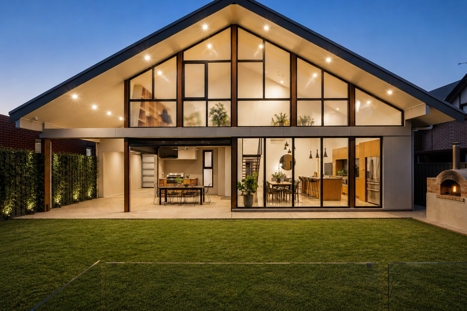 Modern two-story house with large glass windows and an open-concept interior, including a kitchen and dining area, seen during twilight with an illuminated outdoor pizza oven on the right and a green lawn in the foreground.