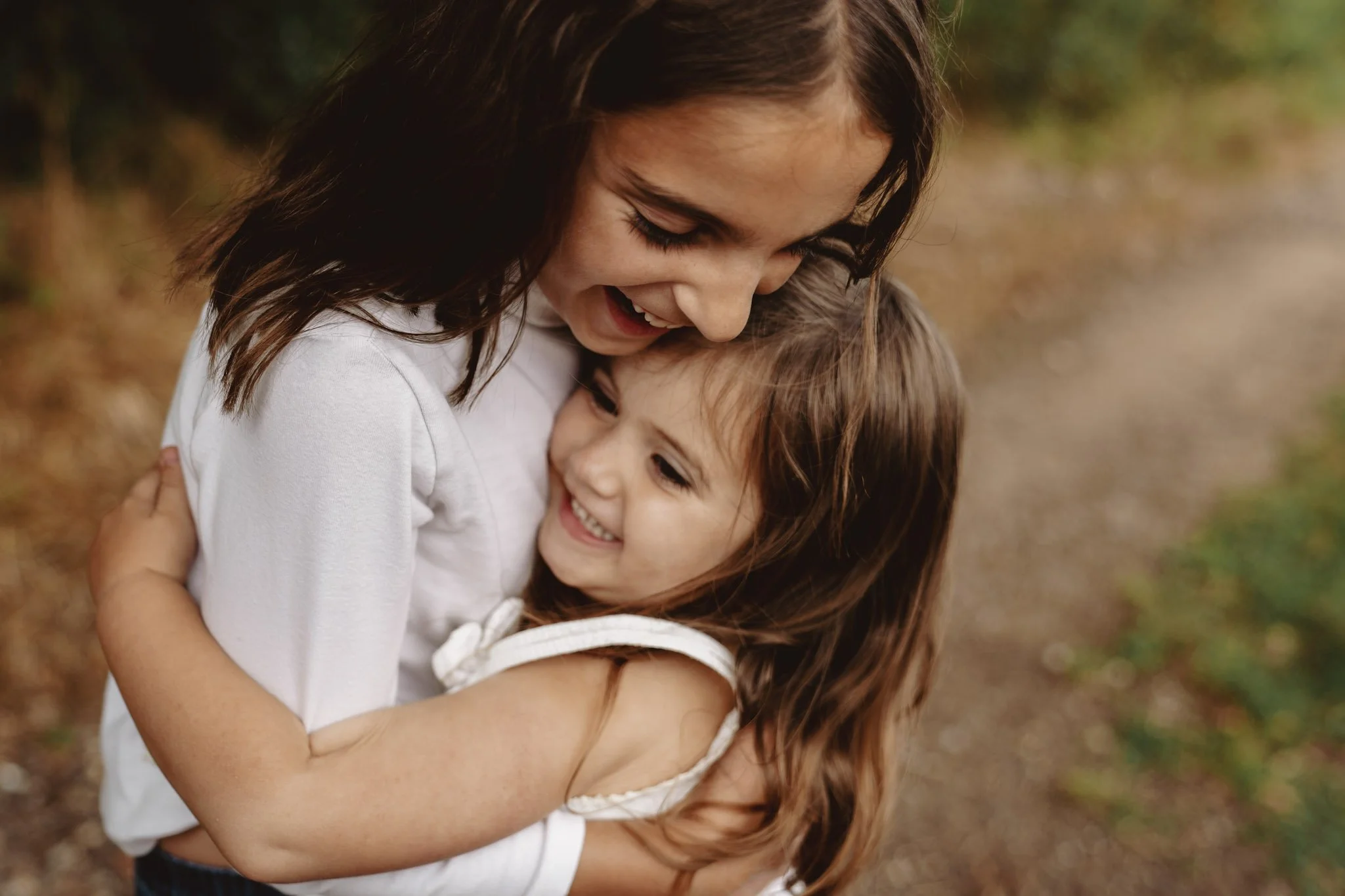 A woman with dark hair smiling and hugging a young girl with long hair outdoors.