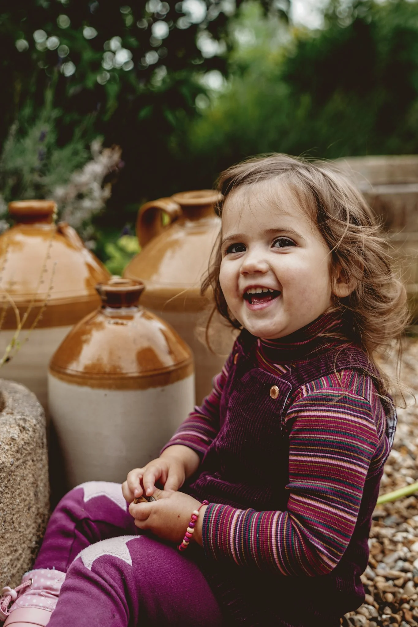 A young girl with curly hair smiling and sitting outdoors, surrounded by large decorative ceramic pots and greenery.