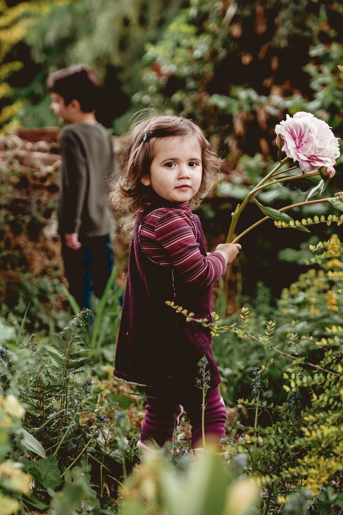 A young girl with curly brown hair and light skin holding a pink peony flower outdoors, with a boy in the background among lush green foliage and flowers.
