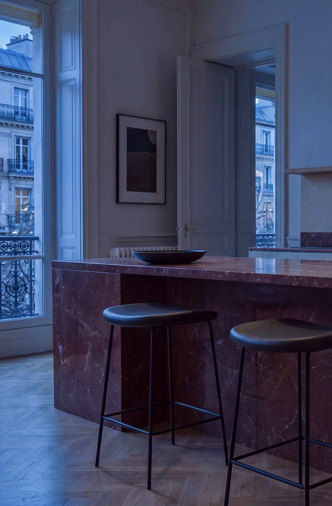 Rendering 3D fotorealistico di una cucina parigina con isola in marmo rosso, sgabelli neri, ampie finestre e luce blu del crepuscolo.