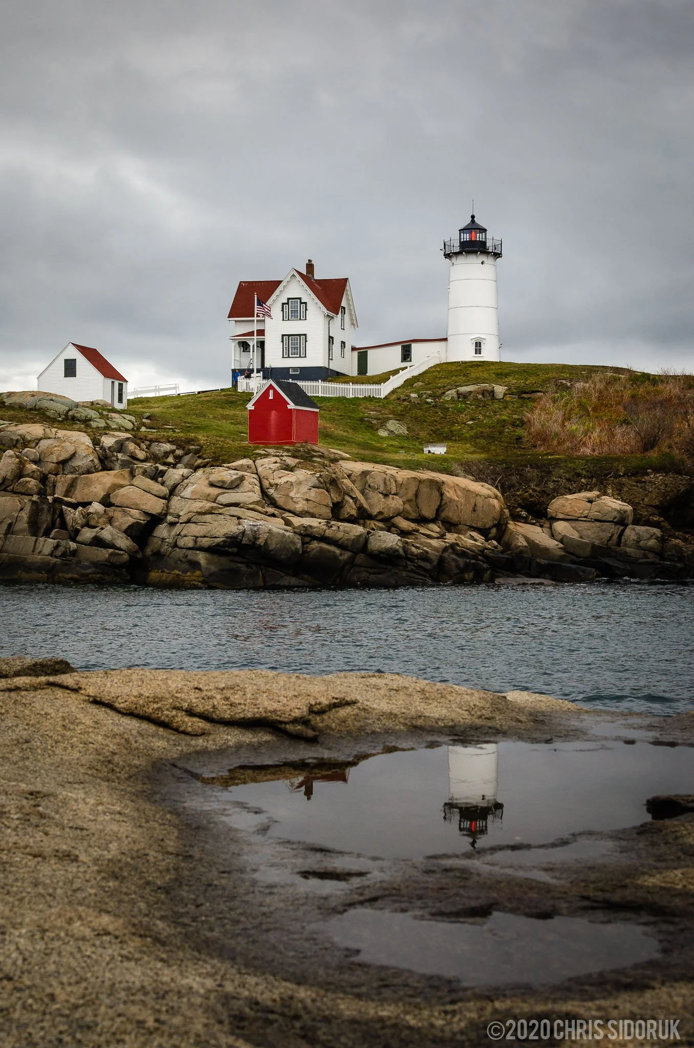 Nubble Lighthouse reflected in a shallow rock pool along the York, Maine coastline with overcast sky and calm water.