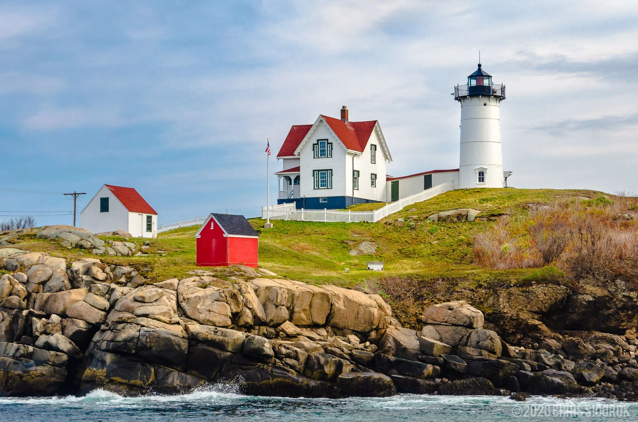 Nubble Lighthouse on a rocky island in York, Maine with surrounding coastal buildings and Atlantic shoreline under soft daylight.