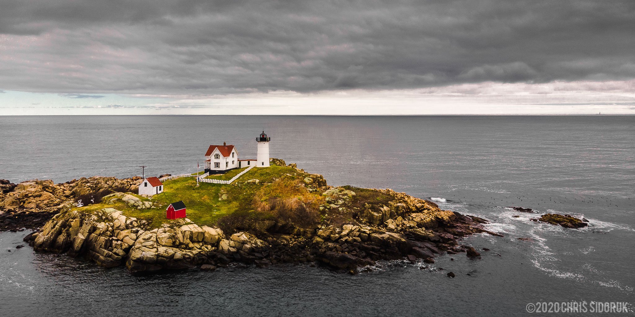 Wide aerial view of Nubble Lighthouse on its small island off York, Maine surrounded by open Atlantic water and rocky coastline.