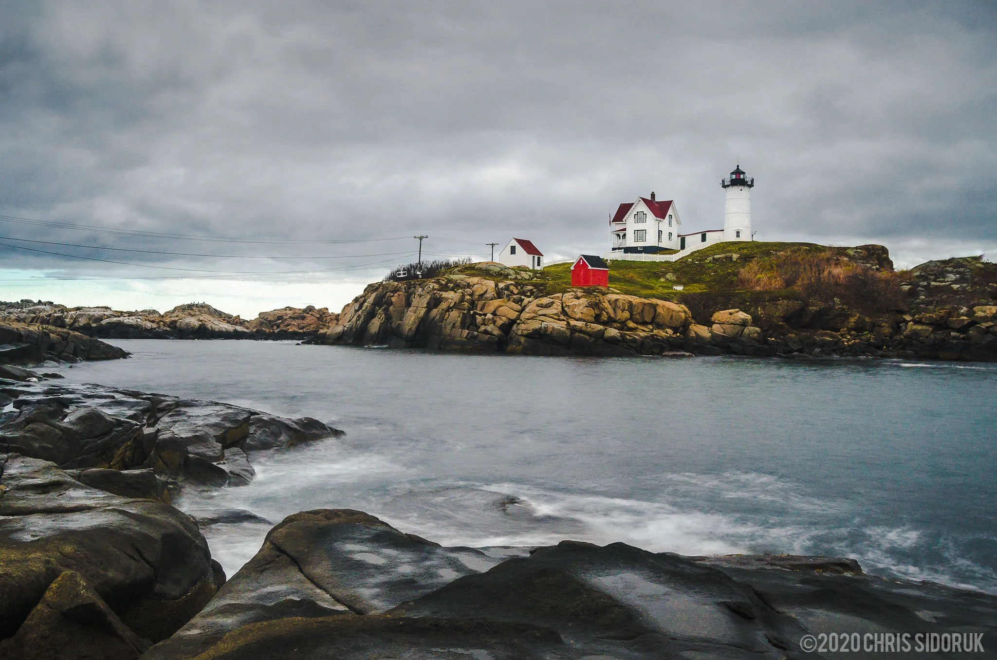 Rocky shoreline in the foreground leading toward Nubble Lighthouse in York, Maine with waves moving along the Atlantic coast.