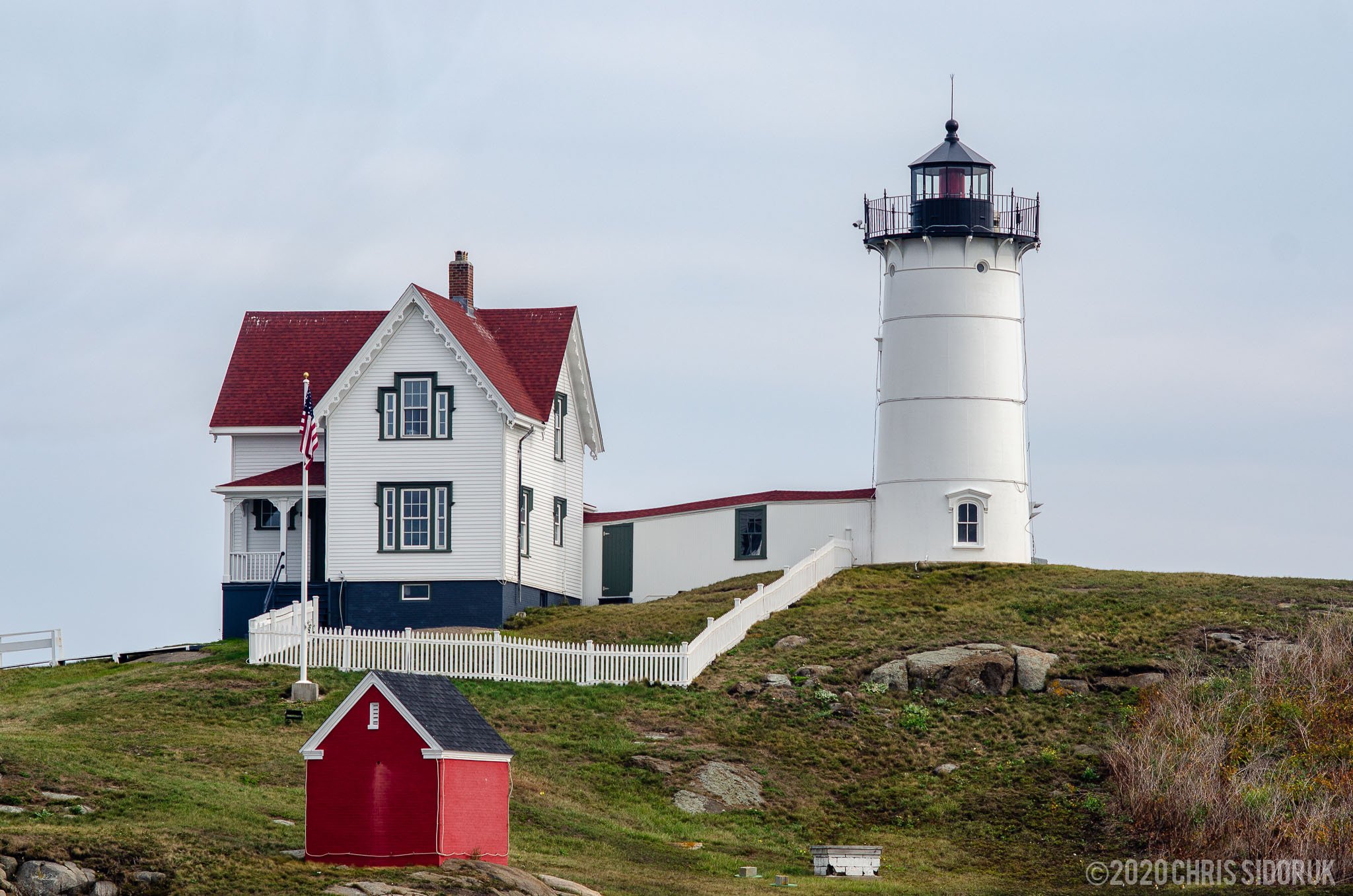 Close view of Nubble Lighthouse and keeper’s house in York, Maine with white fencing and rocky coastal terrain.