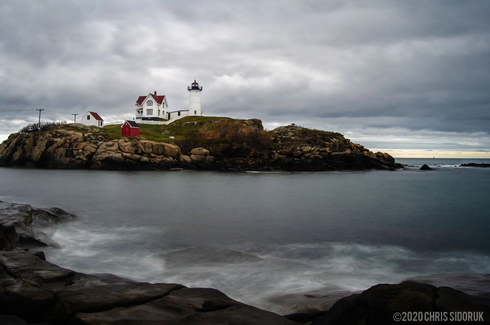 Nubble Lighthouse in York, Maine under dark clouds with long exposure water smoothing along the rocky Atlantic shoreline.