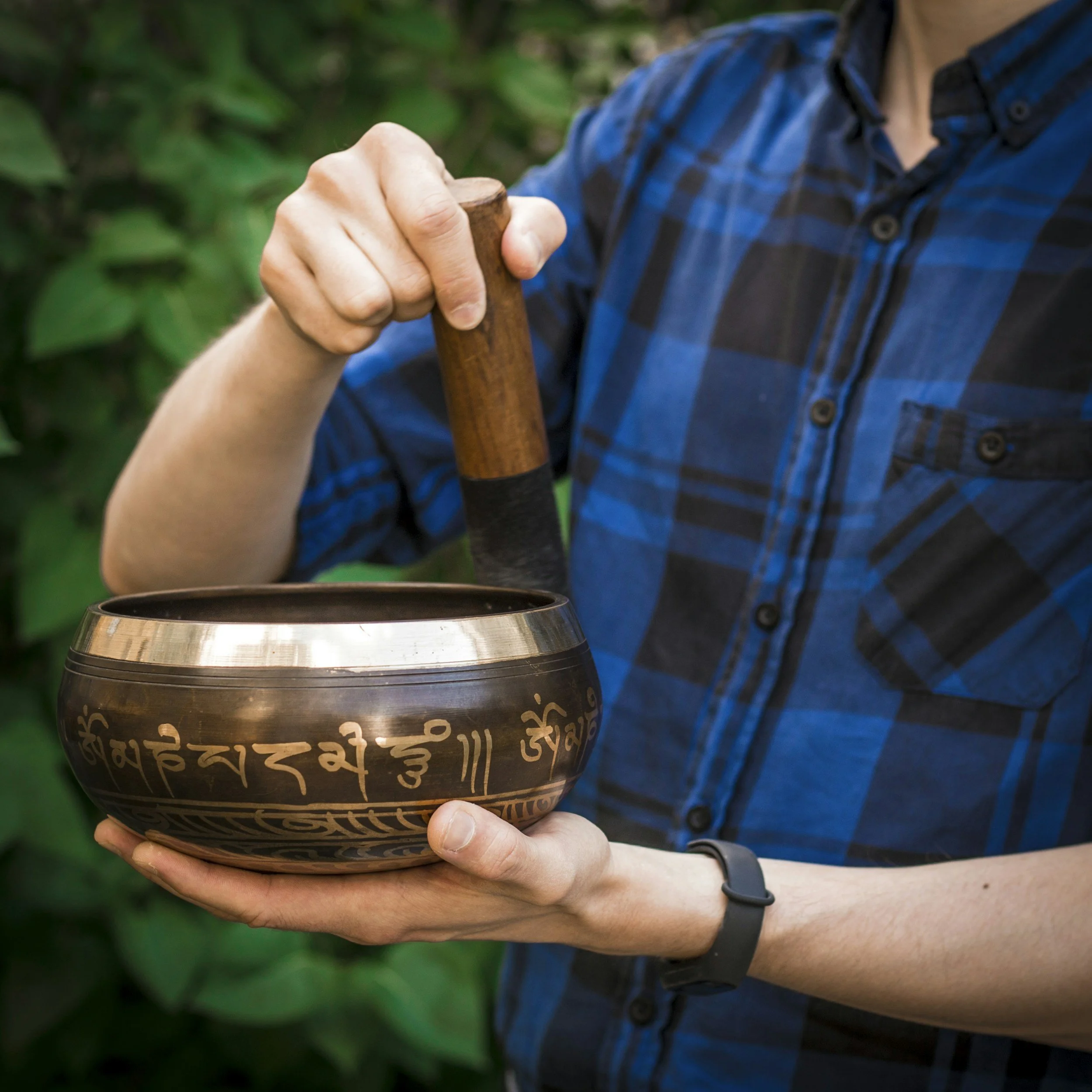 Person holding a Tibetan singing bowl with Tibetan script, using a mallet to strike it, outdoors with green foliage in the background.