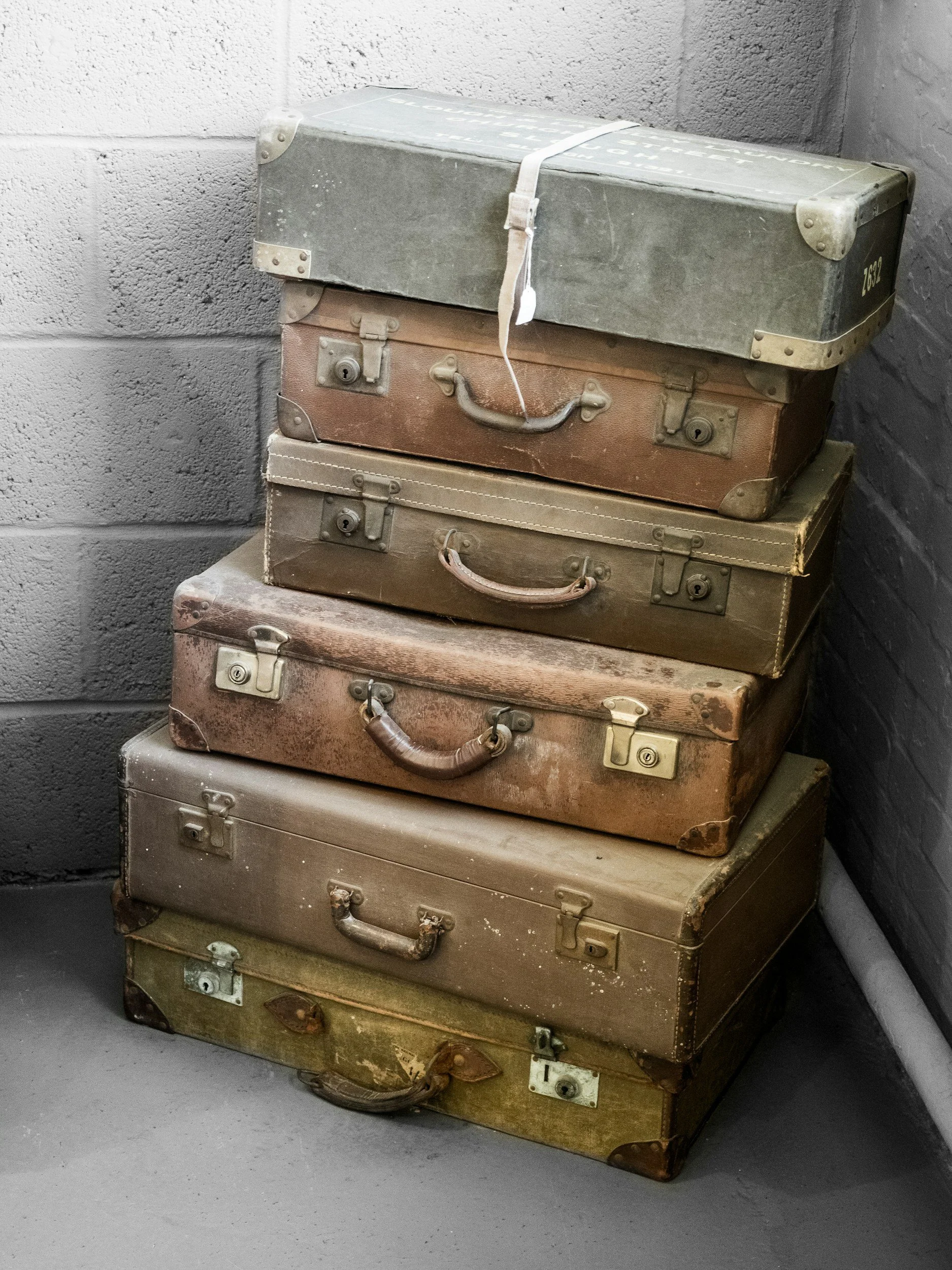 Stack of five vintage suitcases in various shades of brown and gray, with worn handles and metal locks, placed against a gray brick wall.