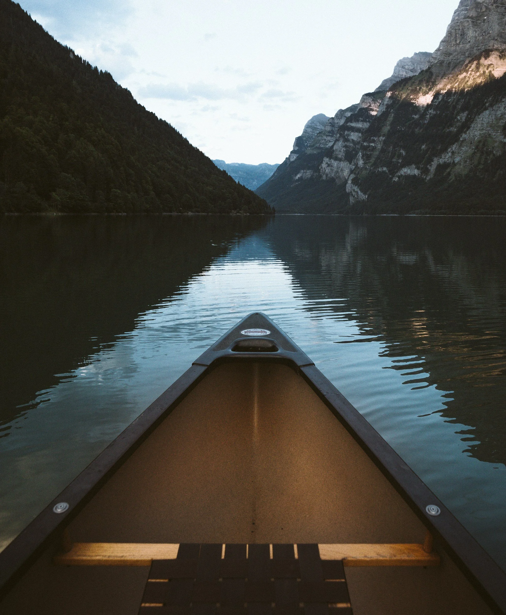 View from a kayak on a calm lake surrounded by mountains with dense tree cover and steep cliffs, under an overcast sky.