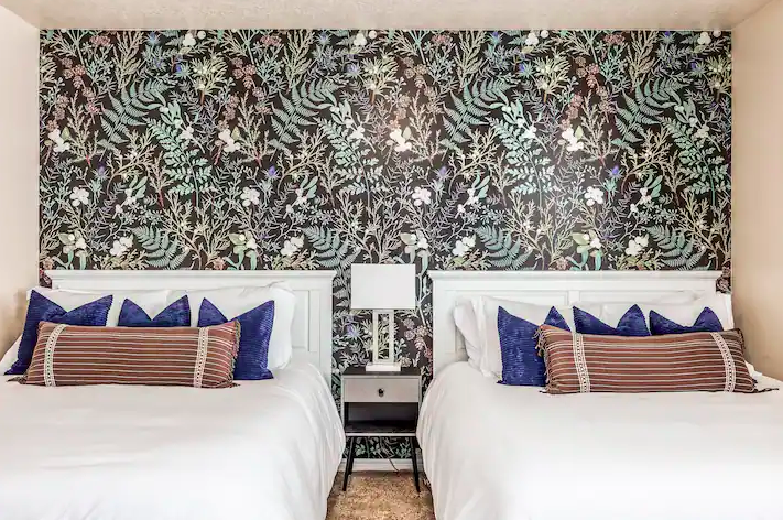 Two queen beds with white linens and decorative pillows, separated by a nightstand with a white lamp, in front of a floral wallpapered wall.