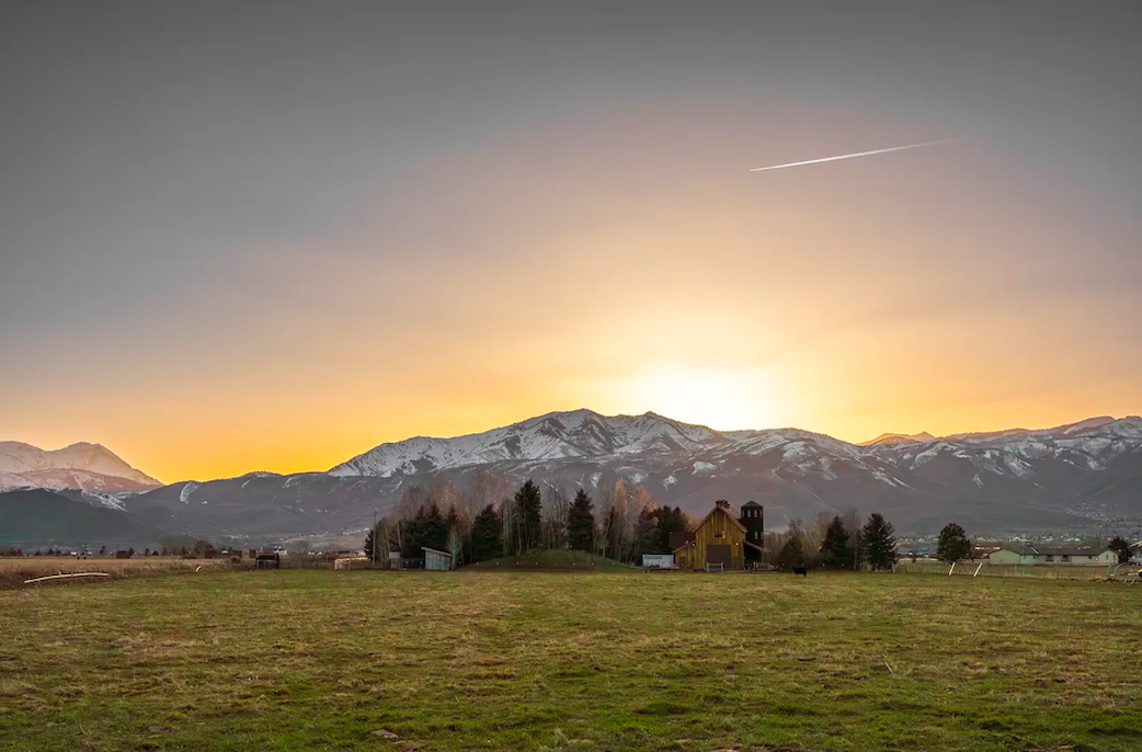 A rural farm landscape at sunset with snow-capped mountains in the background. There are a few farm buildings and trees in the foreground.