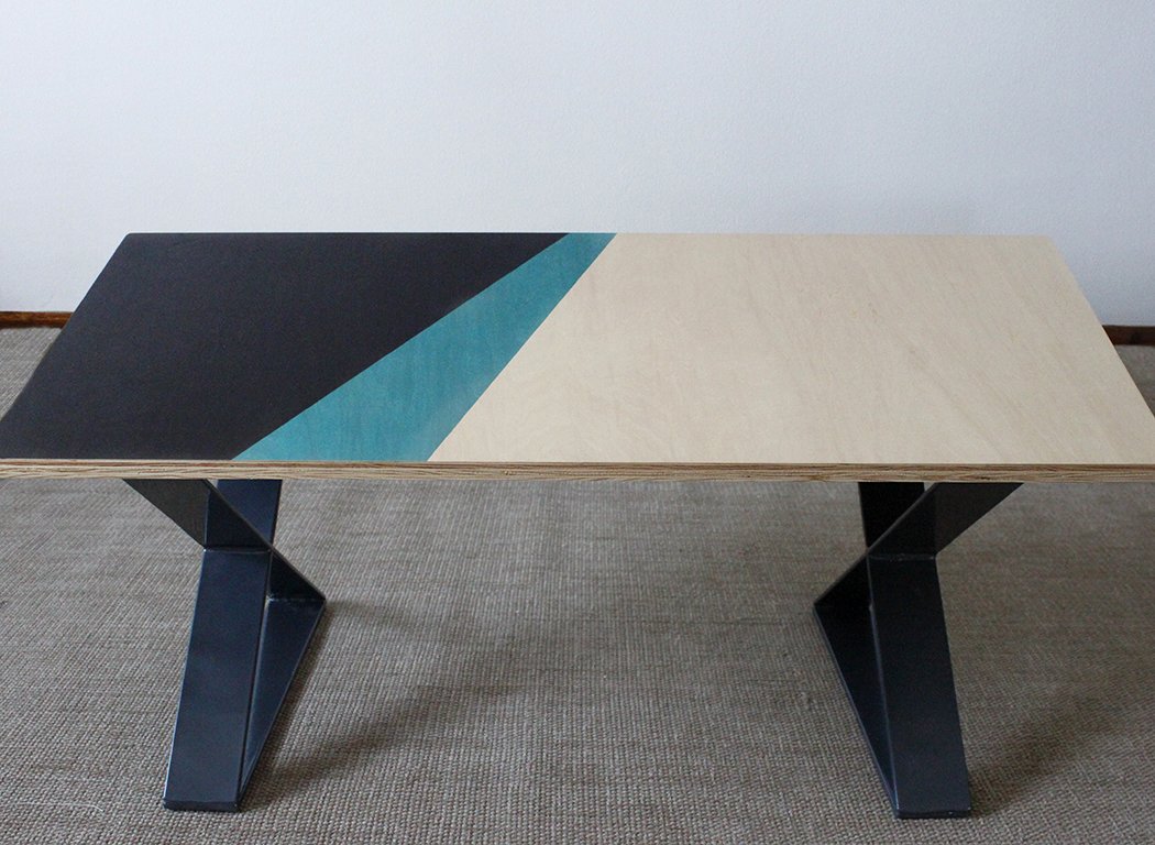 Modern coffee table with geometric design featuring black, teal, and natural wood colors on a gray carpet.