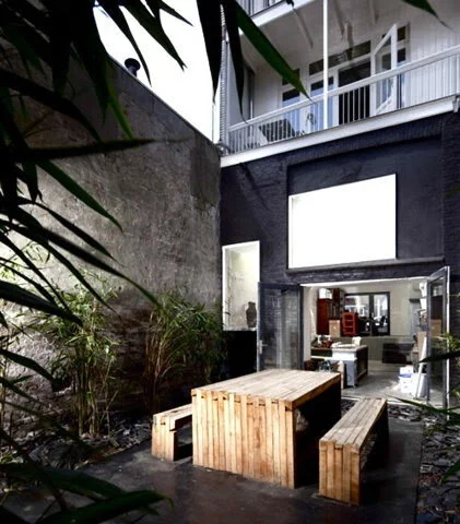 Outdoor patio with wooden furniture, bordered by a high concrete wall and plants, leading into a modern building with glass doors.