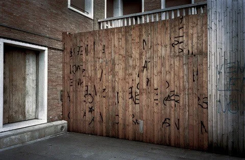 Wooden fence with black symbols and letters, surrounded by brick buildings.