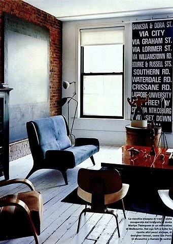Modern living room with brick wall, blue sofa, wooden chairs, white painted flooring, and large bus roll art on wall.