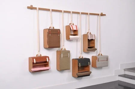 Wall-mounted shelves resembling suitcases hang on a white wall. The shelves are made of wood and fabric, featuring various colors like brown, gray, and pink. They are suspended by straps from a horizontal wooden rod.