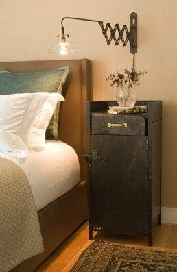 Bedroom scene with a modern industrial nightstand, featuring a metal cabinet with a drawer. A scissor-arm wall lamp is mounted above, extending to illuminate the area. A plush bed is partially visible, adorned with pillows and a beige quilt. A small 