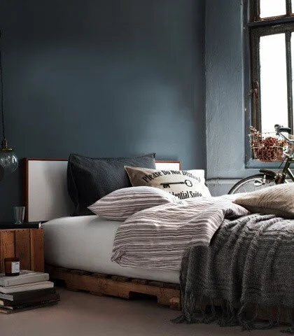 Cozy bedroom with pallet bed, striped and gray bedding, pillows, a wooden nightstand, and a bicycle by the window.