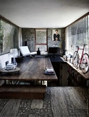 Modern interior with large wooden table, white lounge chair, bicycle on display, and bookshelves, stone walls, and large windows.