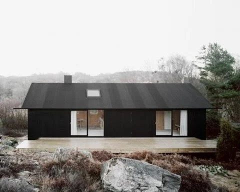 Modern minimalist black house with large glass windows, surrounded by rocky landscape and sparse vegetation.