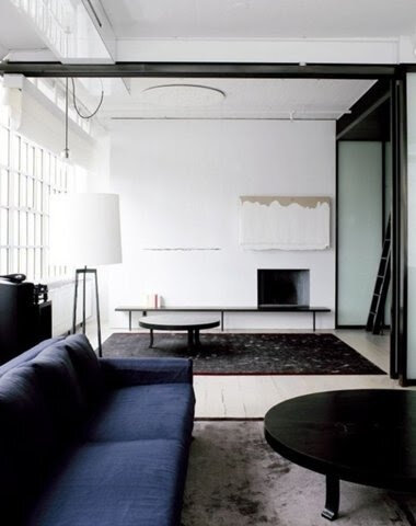 Modern living room with a dark blue sofa, round black coffee table, large floor lamp, and minimalist decor. There's a black rug, abstract painting, and large windows on the side. The room has sliding glass doors.