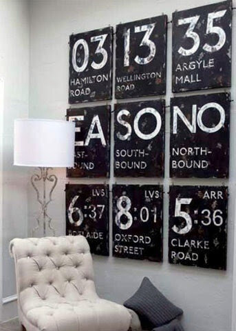 Wall art with nine vintage-style transit signs featuring destinations and numbers, next to a tall lamp and tufted chair in a modern interior.
