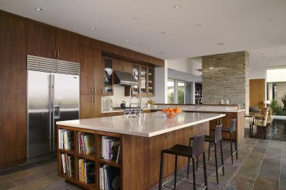 Modern kitchen with wooden cabinets, large island with bar stools, built-in refrigerator, bookshelves, and stone floor.