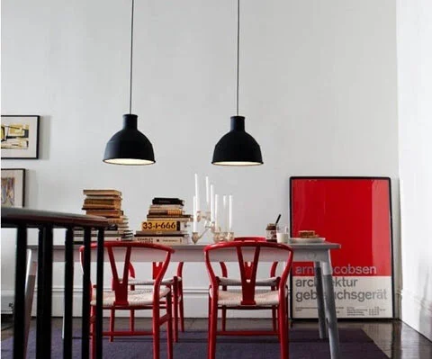 Dining area with red chairs and modern decor, featuring black pendant lights, a stack of books on the table, and a large red and white art piece leaning against the wall.