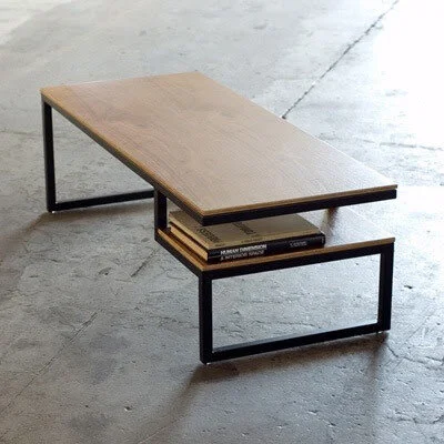 Modern wooden coffee table with black metal frame and a lower shelf holding books, placed on a concrete floor.