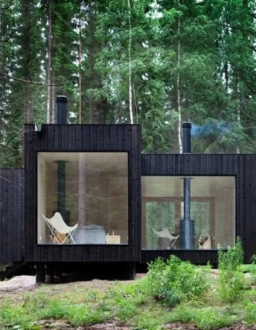 Modern black cabin with large windows in a forest setting, featuring a butterfly chair visible inside.
