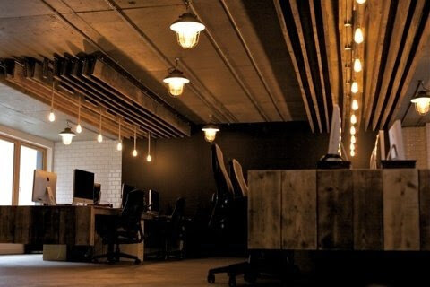 Modern office space with wooden decor and industrial lighting, featuring desks, chairs, and computer monitors.