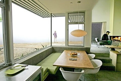 Mid-century modern interior with dining area, green cushions, large windows, and pendant lamp. Background shows a foggy outdoor view and a person by a fireplace.