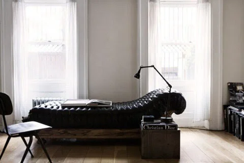 Minimalist room with a black leather chaise lounge, two large windows with white curtains, and a side table holding books and a lamp.