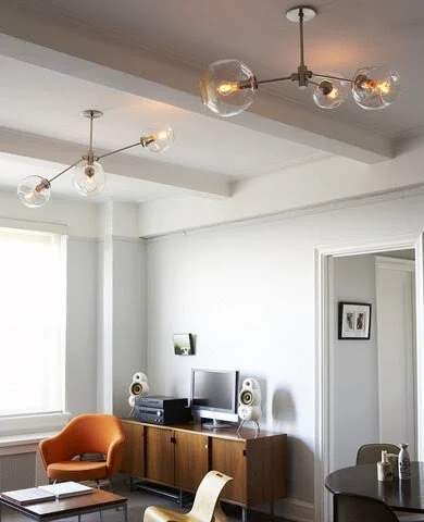 Modern living room with two hanging glass globe chandeliers, a wooden sideboard, computer monitor, speakers, an orange armchair, and a small round dining table with chairs.