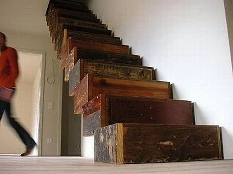 A staircase made of stacked wooden drawers attached to a wall with a blurred person walking by.