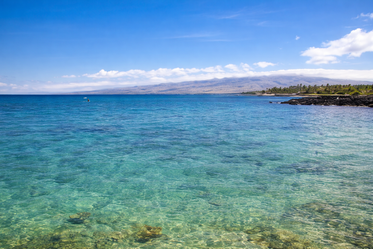 Clear blue ocean water with a rocky shoreline on the right and distant mountains on the horizon under a bright blue sky with some white clouds.