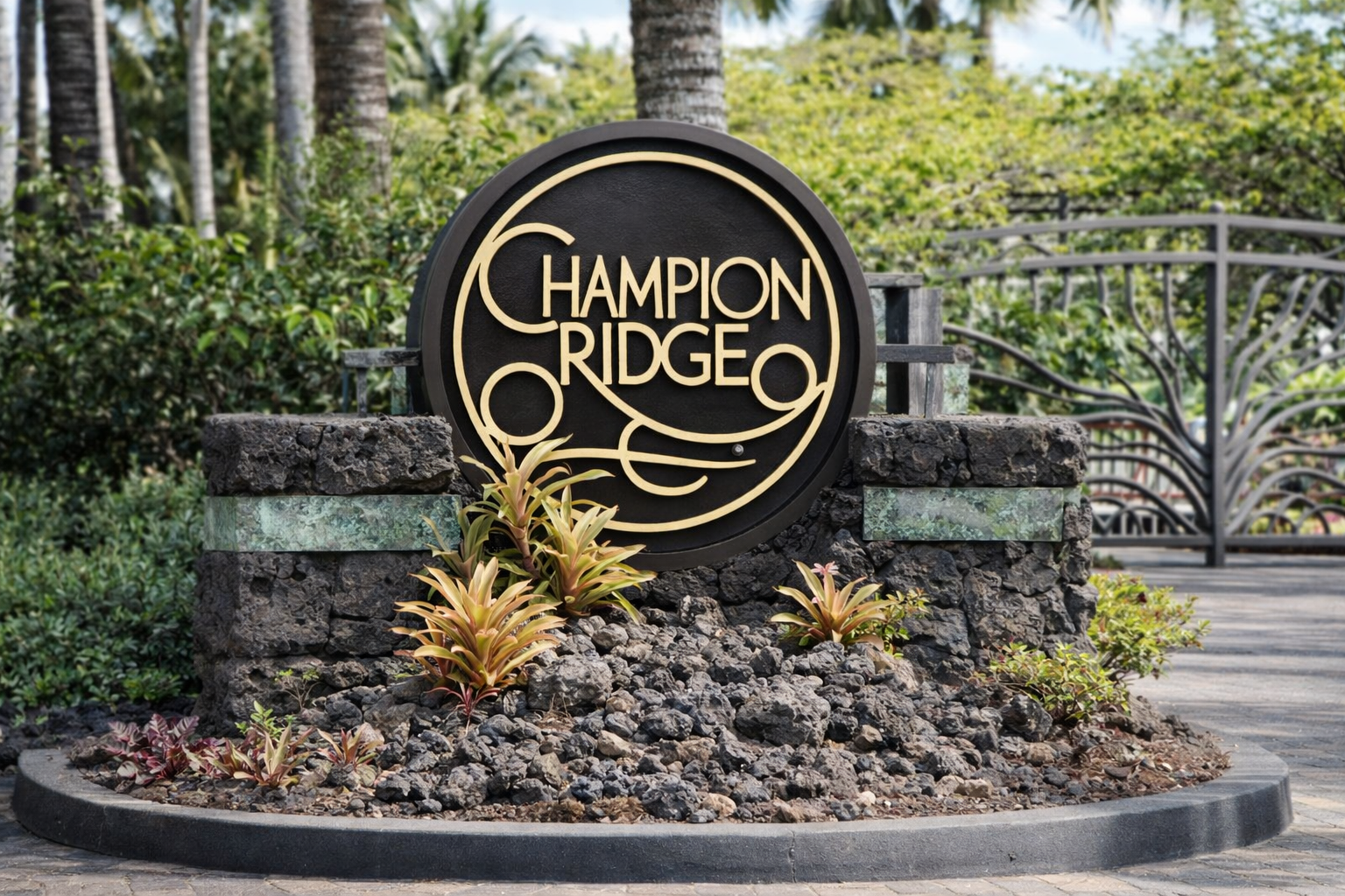 Tropical park entrance with palm trees, bushes, a paved walkway, and a circular flower bed at entrance of Champion Ridge Neighborhood.