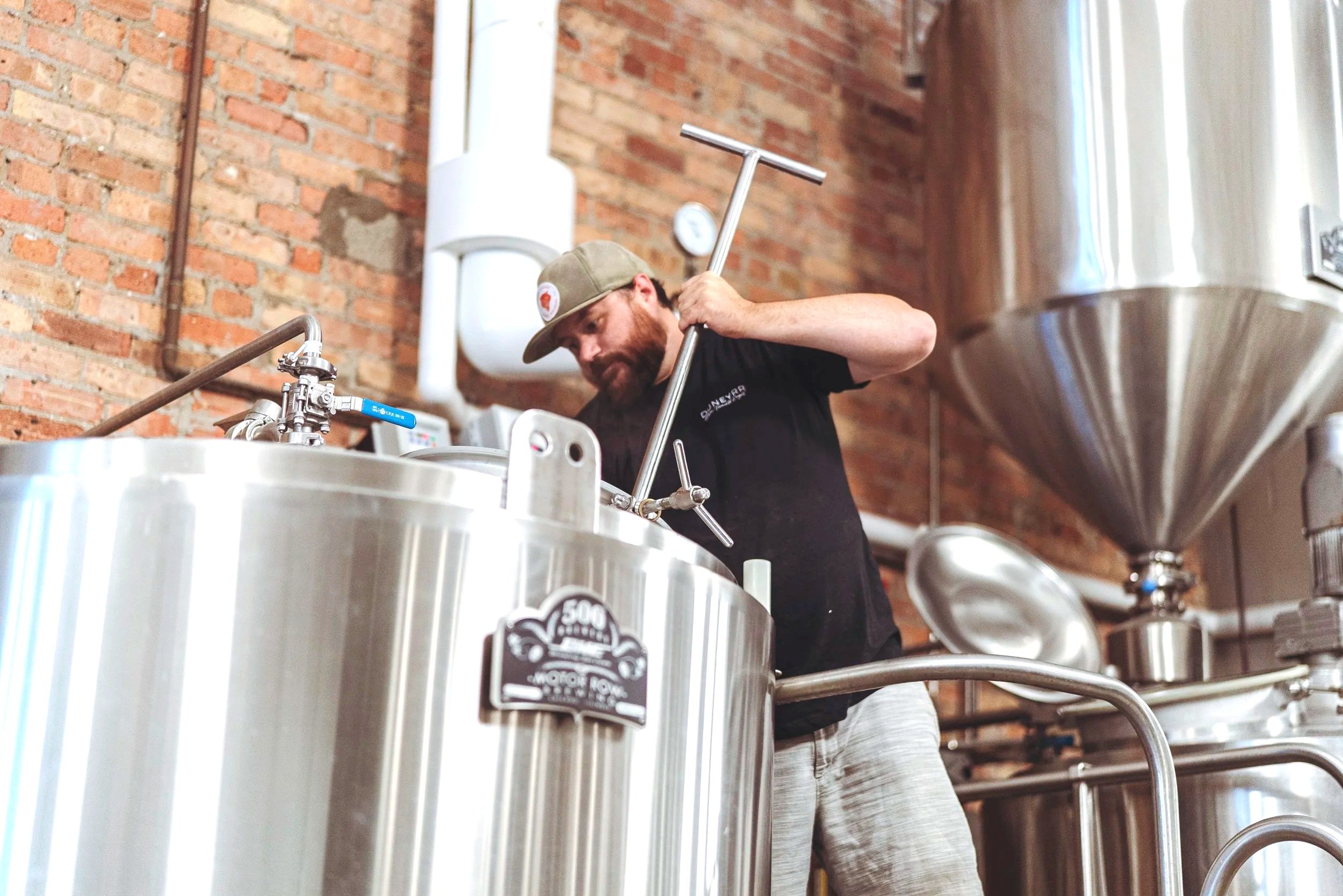 A man is working inside a brewery, stirring a large stainless steel tank with a long-handled tool. The brewery has exposed brick walls and various brewing equipment.